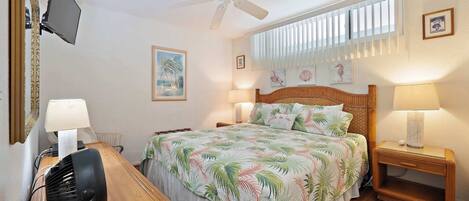 A bedroom with a bed, two bedside tables, a ceiling fan, a TV mounted on the wall, and a window with blinds.
