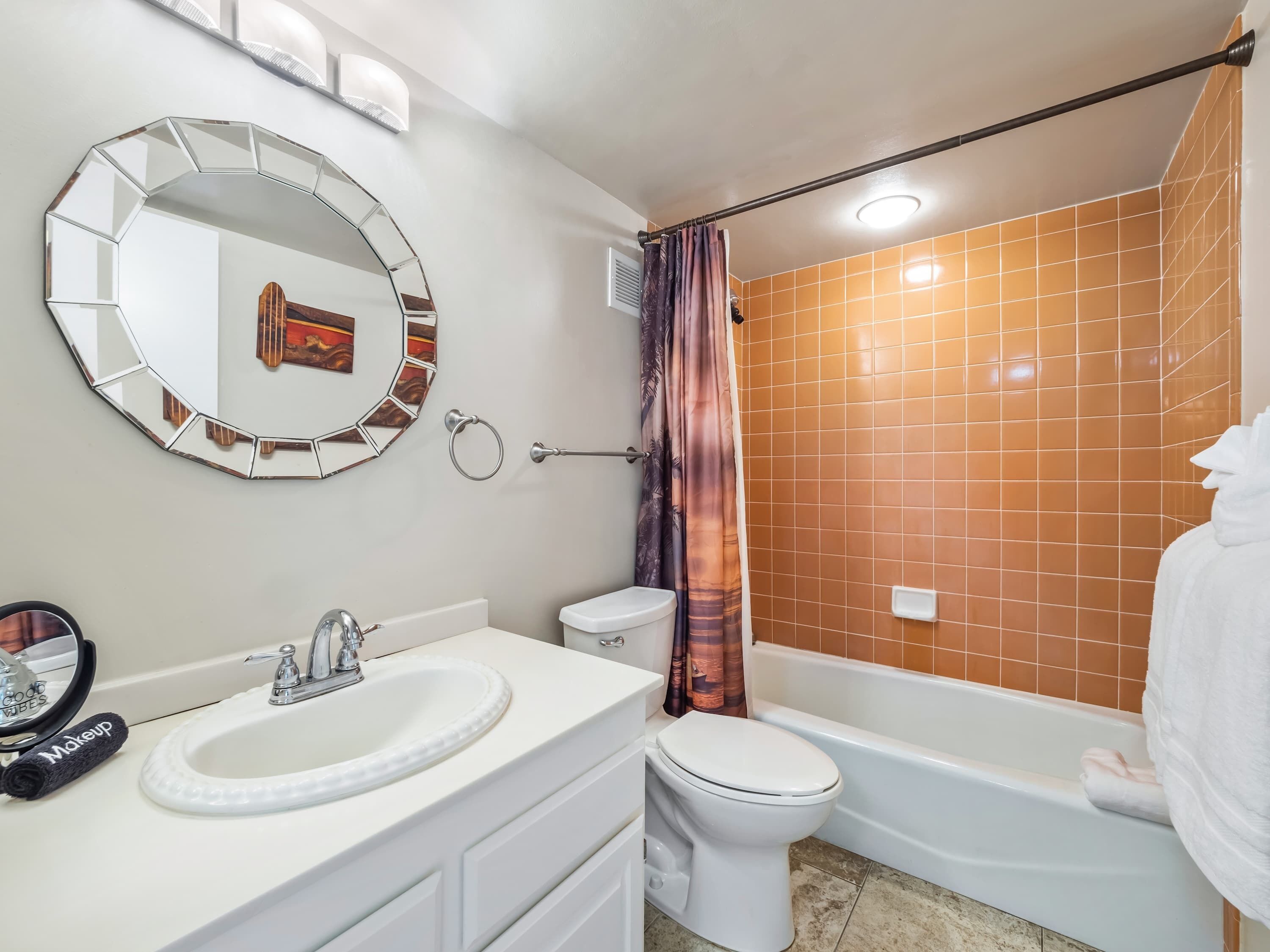 Een badkamer met een witte wastafel, toilet en badkuip. Er is een spiegel met een geometrisch ontwerp aan de muur, en een handdoekhouder met een handdoek.