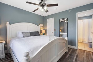 A bedroom with a white bed, a ceiling fan, a nightstand, a lamp, and a door leading to a bathroom. - Eagle's Nest at The Club- Chic 2BR Golf View Condo (Branson)