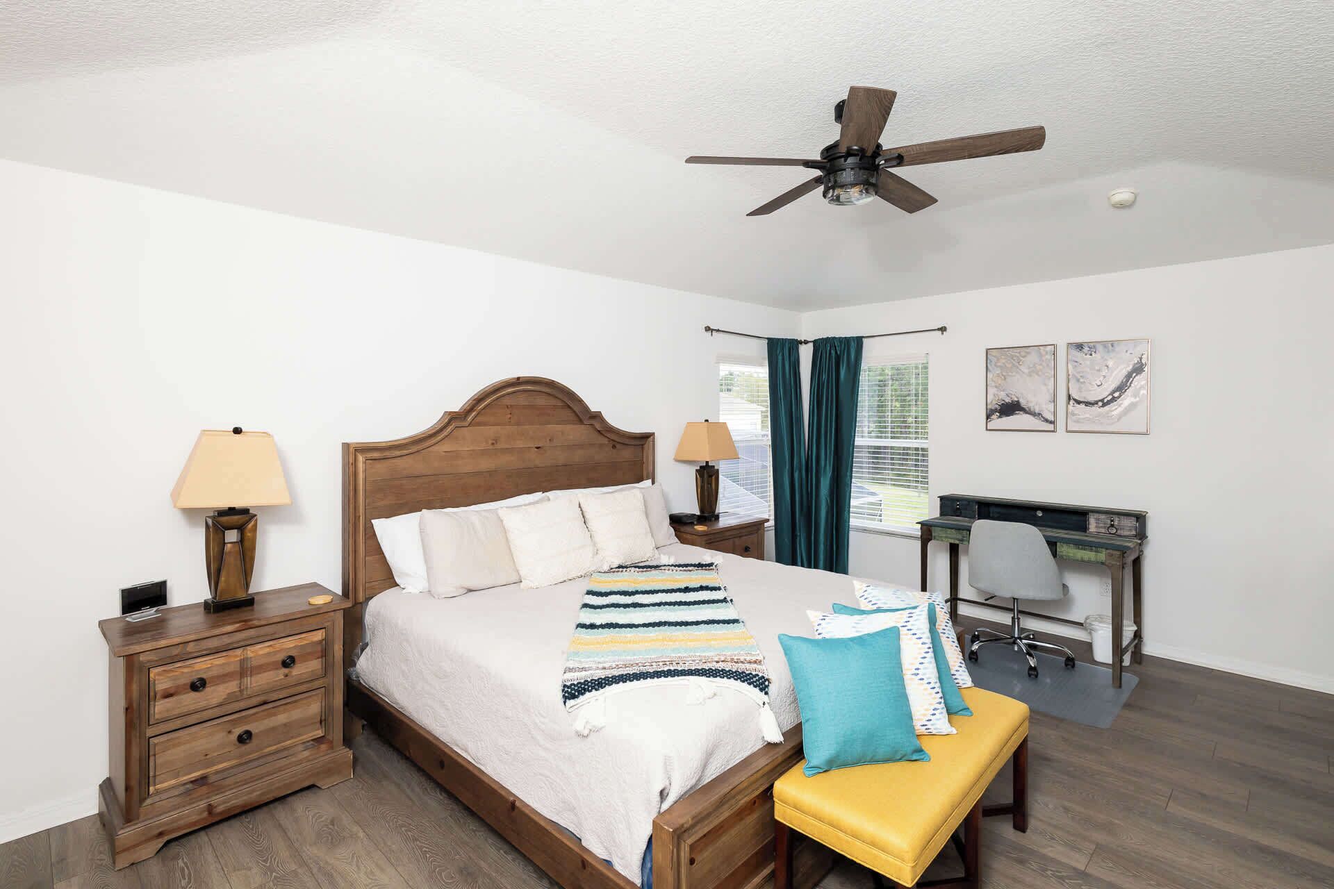 A bedroom with a wooden bed, two bedside tables with lamps, a desk with a chair, and a ceiling fan.