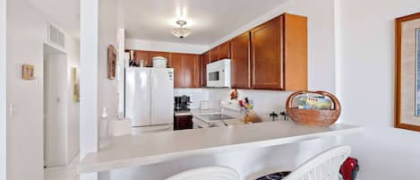 A modern kitchen with wooden cabinets, a white refrigerator, a microwave, and a coffee maker. There are two wicker chairs at the counter.