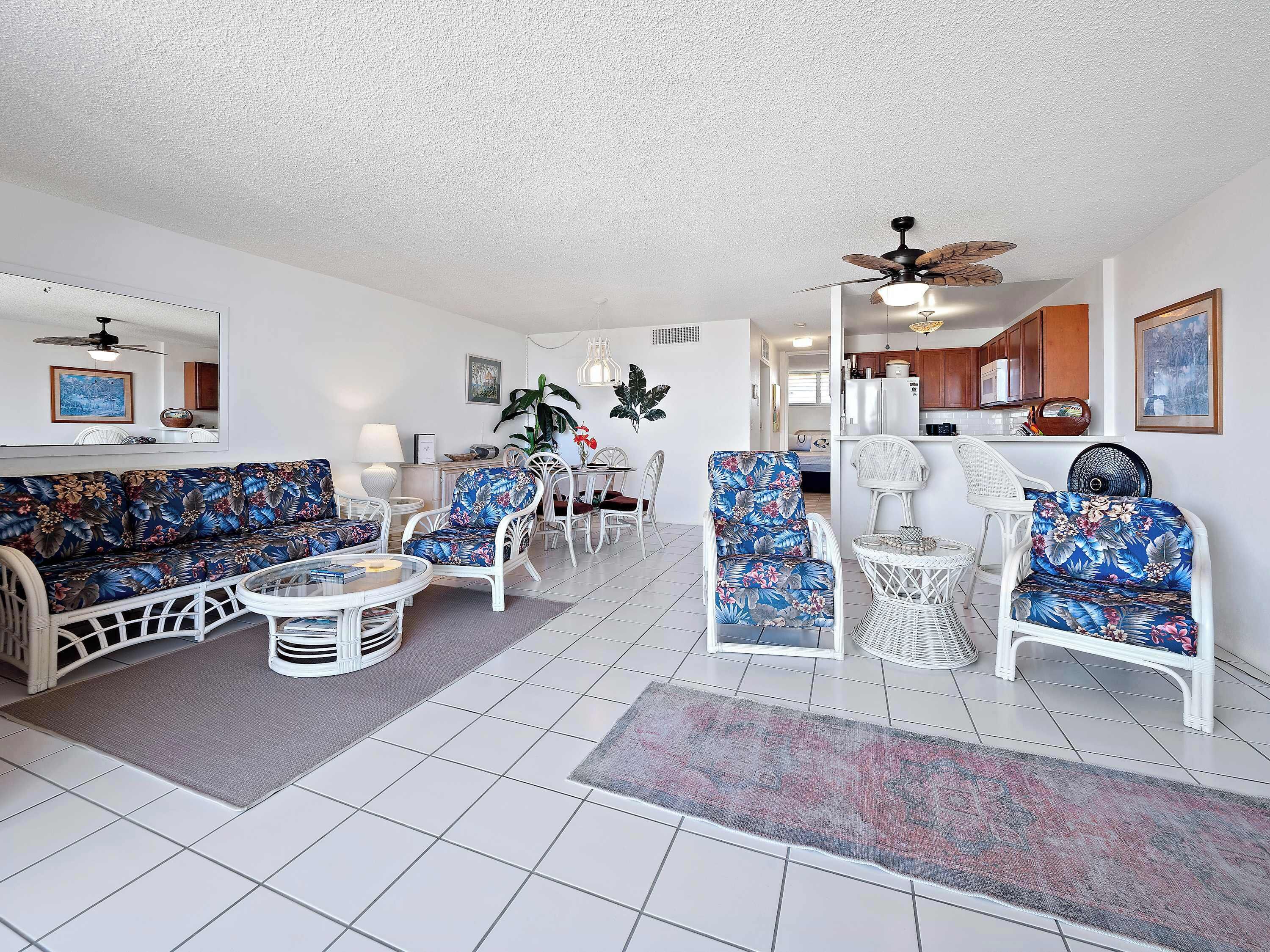 A spacious living area with a sofa, armchairs, and a coffee table. There is a ceiling fan and a dining area visible in the background.