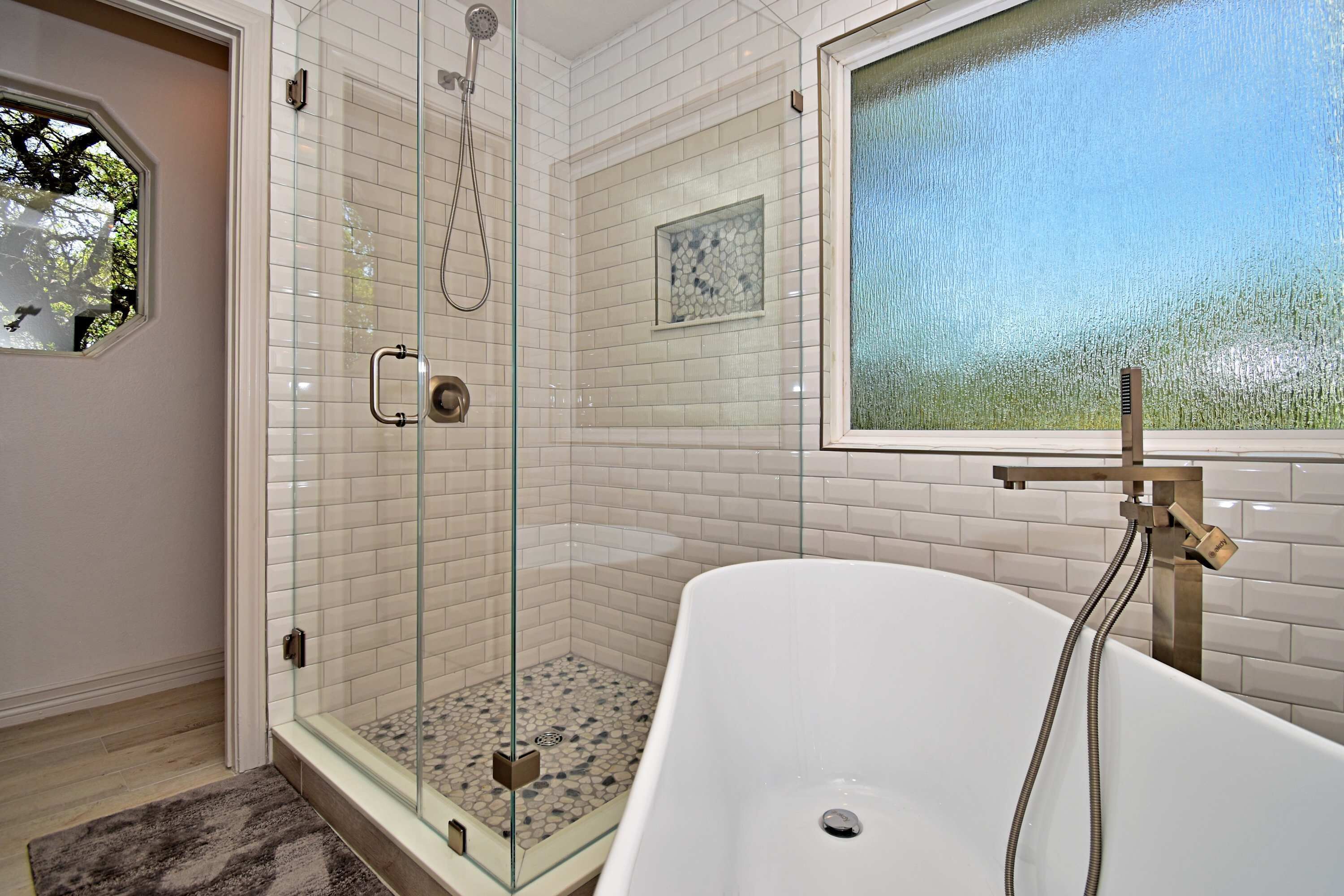 A modern bathroom with a glass-enclosed shower, a freestanding bathtub, and a window with a view of trees.