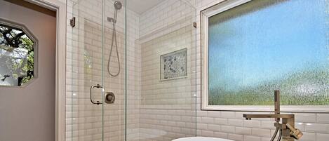 A modern bathroom with a glass-enclosed shower, a freestanding bathtub, and a window with a view of trees.