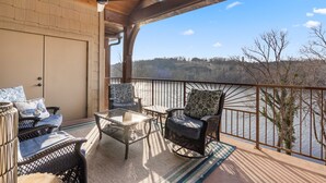 A covered outdoor seating area with wicker furniture and a glass table, overlooking a body of water and trees. - Penthouse 7 on Taneycomo - Chic Waterfront Condo! (Branson)
