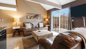 Ein gemütliches Wohnzimmer mit einer braunen Ledersofa, einem gemusterten Hocker und einem großen Fenster mit Blick auf Bäume.