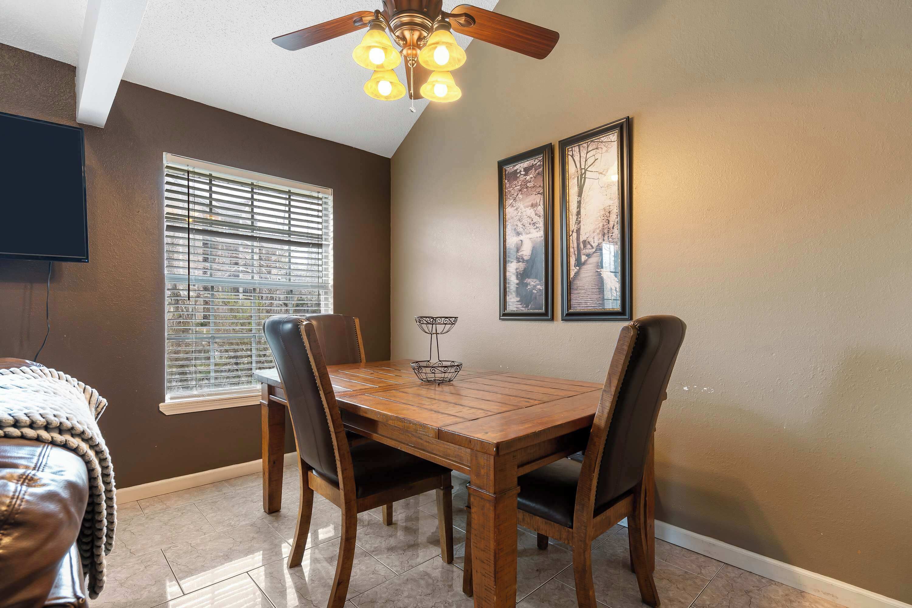 A dining area with a wooden table and chairs, a ceiling fan, a window with blinds, and framed artwork on the wall.