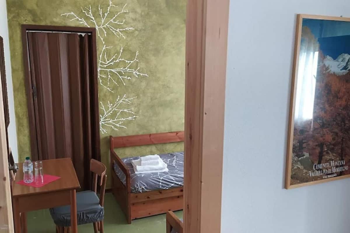 A small room with a wooden bed, a desk with a chair, a door, and a wall with a framed picture.