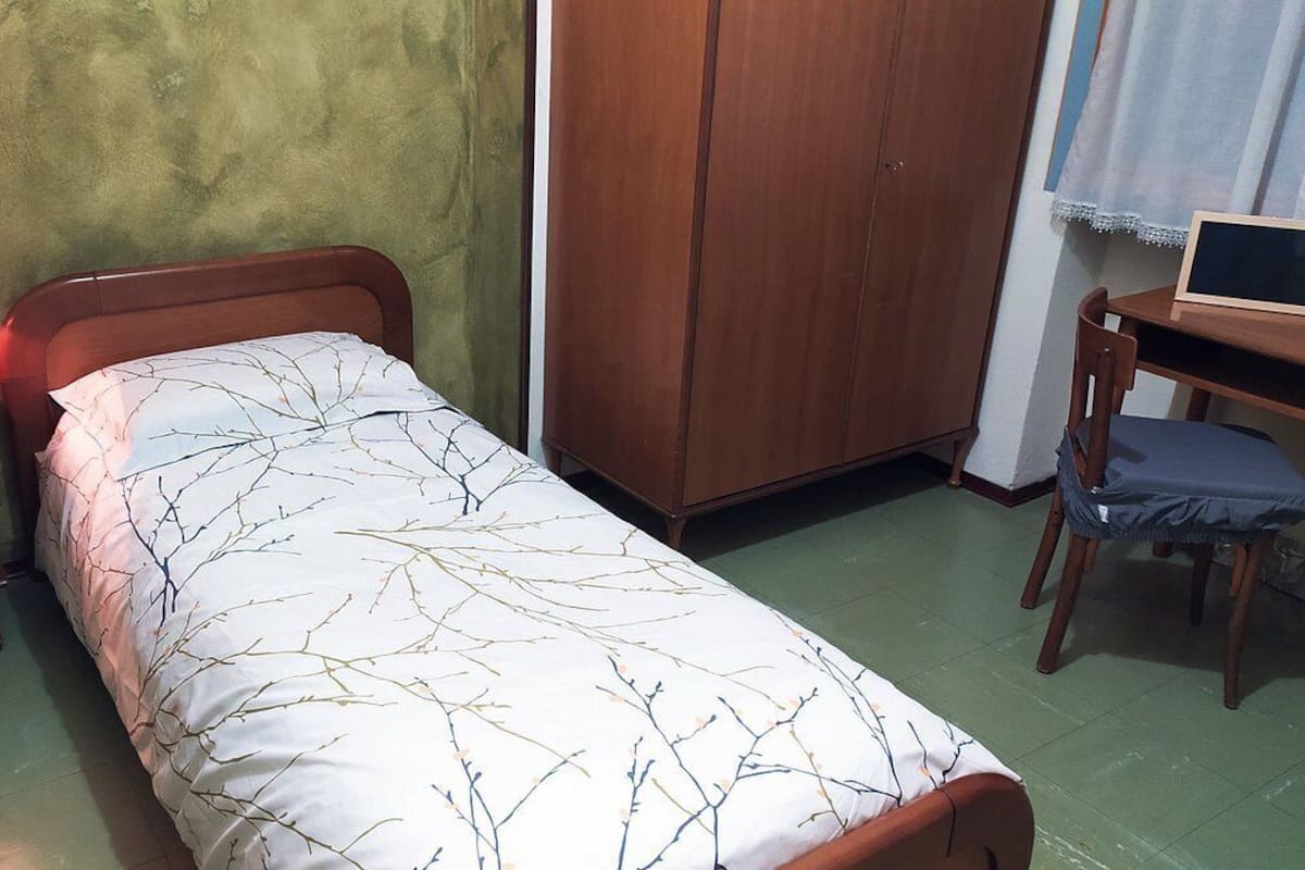 A small bedroom with a single bed, a nightstand with a lamp, a wooden wardrobe, and a desk with a chair.