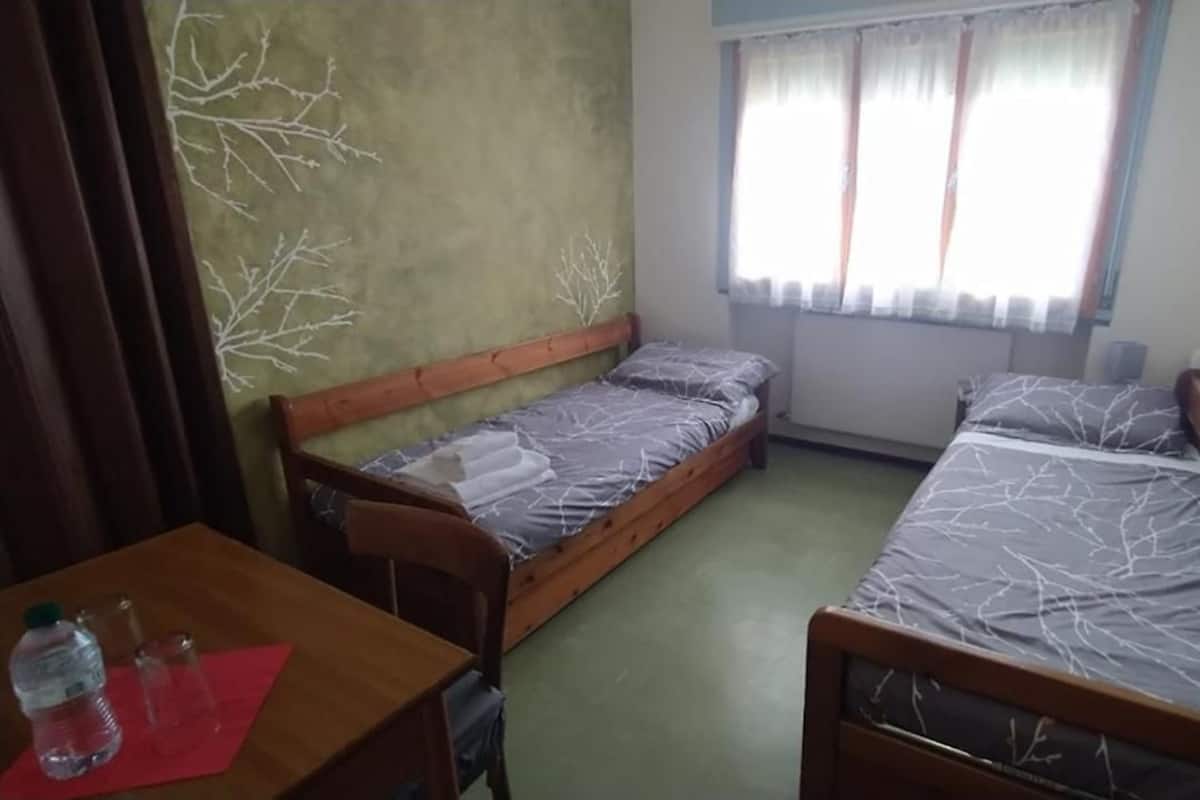 A small, simple room with two wooden beds, a table with a bottle and glasses, and a window with curtains.