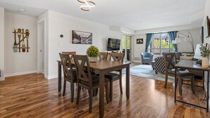 Een ruime woonkamer met een houten eettafel, zachte zithoek, een televisie en een groot raam met gordijnen.