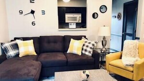 Un salon avec un canapé marron foncé, un fauteuil jaune et une table basse. Un four à micro-ondes est fixé au mur, au-dessus du canapé.