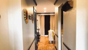 Un couloir étroit au plancher en bois, une chaise recouverte d’une housse blanche et une porte au fond.