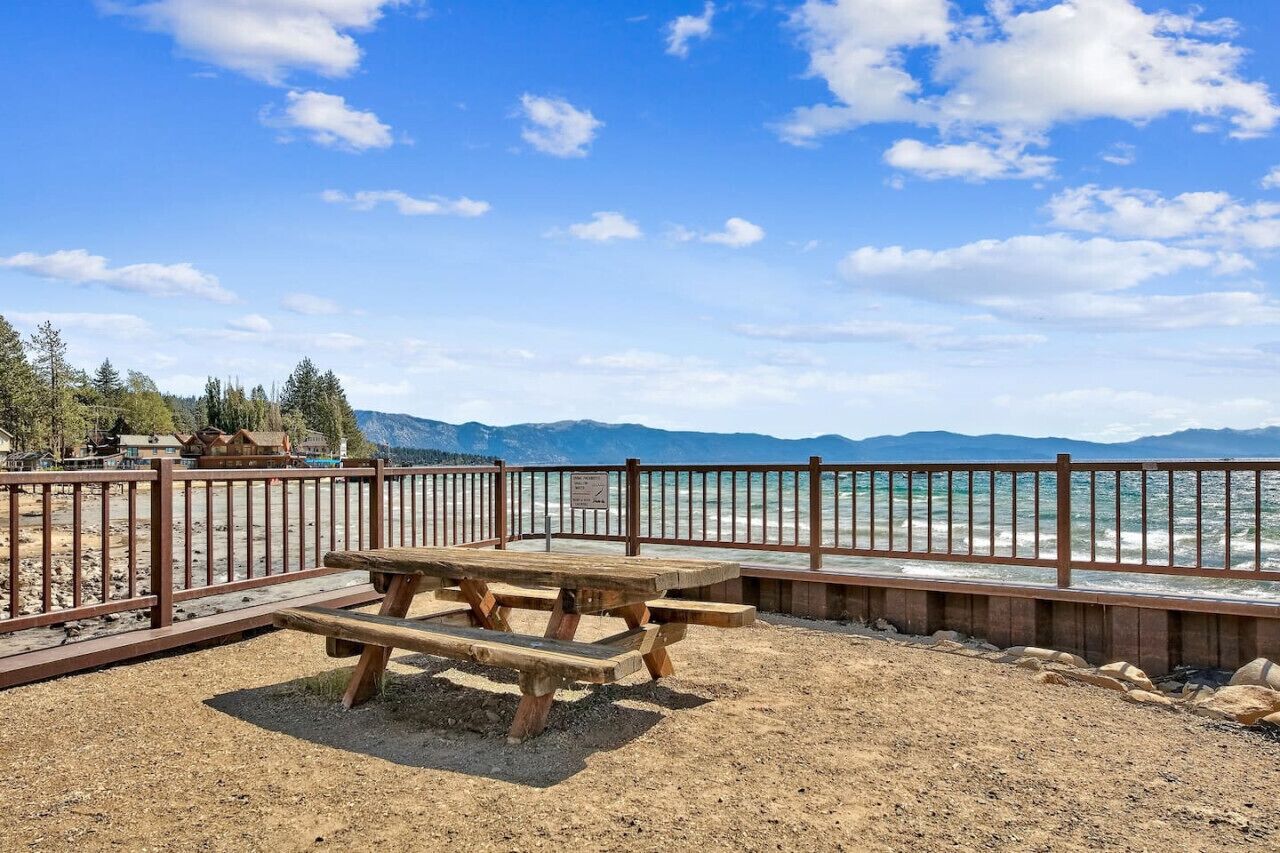 Une table de pique-nique en bois avec des bancs, une clôture en métal et une vue sur l’océan et les montagnes.