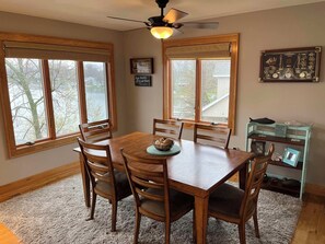 A dining area with a wooden table and chairs, large windows with blinds, a ceiling fan, and a small shelf with decorative items. - Lakefront Home w/ Sunset Views. New to Rental Market! Pontoon Rental Available. (Plainwell)