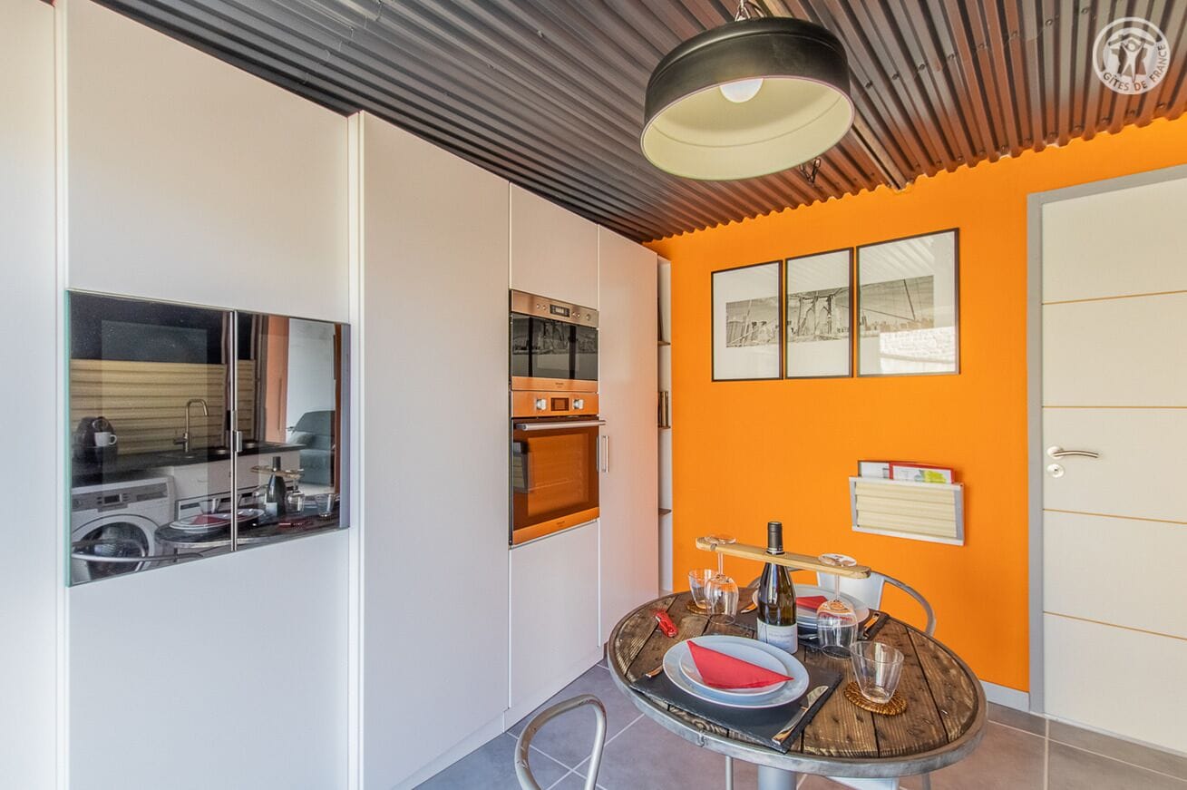 Une cuisine compacte dotée d’appareils électroménagers modernes, un coin repas avec une table en bois et un mur d’accent d’une couleur orange vive.