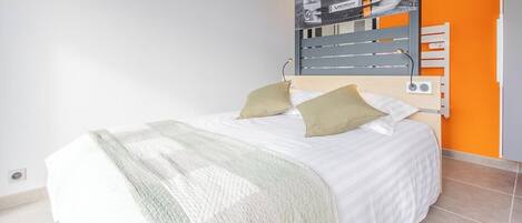 A bed with a white bedspread and two pillows, a headboard with a mounted picture, and a wall with an orange accent.