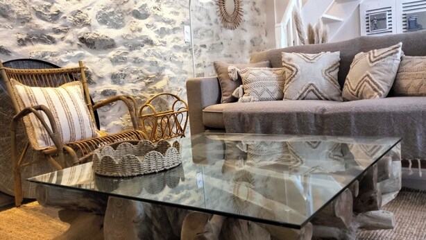 Un salon avec un mur en pierre, un fauteuil en osier, une table basse en verre et un canapé gris agrémenté de coussins à motifs.