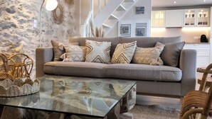 Un salon moderne avec un canapé gris, des coussins à motifs, une table basse en verre, des paniers en osier et une cuisine en arrière-plan.