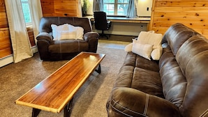 Una sala de estar acogedora con un sofá de cuero marrón, una mesa de centro de madera y un sillón de cuero. Se ve un escritorio con una computadora y una silla al fondo.