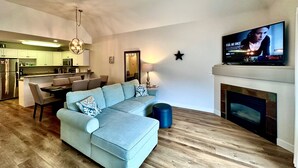 Una sala de estar moderna con un sofá seccional azul claro, un comedor con mesa y sillas, una cocina al fondo, una chimenea y un televisor que muestra un programa.