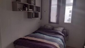 Ein kleiner, einfach eingerichteter Raum mit einem Bett, einem Fenster und fest eingebauten Regalen.