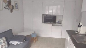 Ein modernes Wohnzimmer mit grauem Sofa, einem kleinen runden Tisch und einer Küchenzeile mit weißen Schränken sowie einem Fernseher.