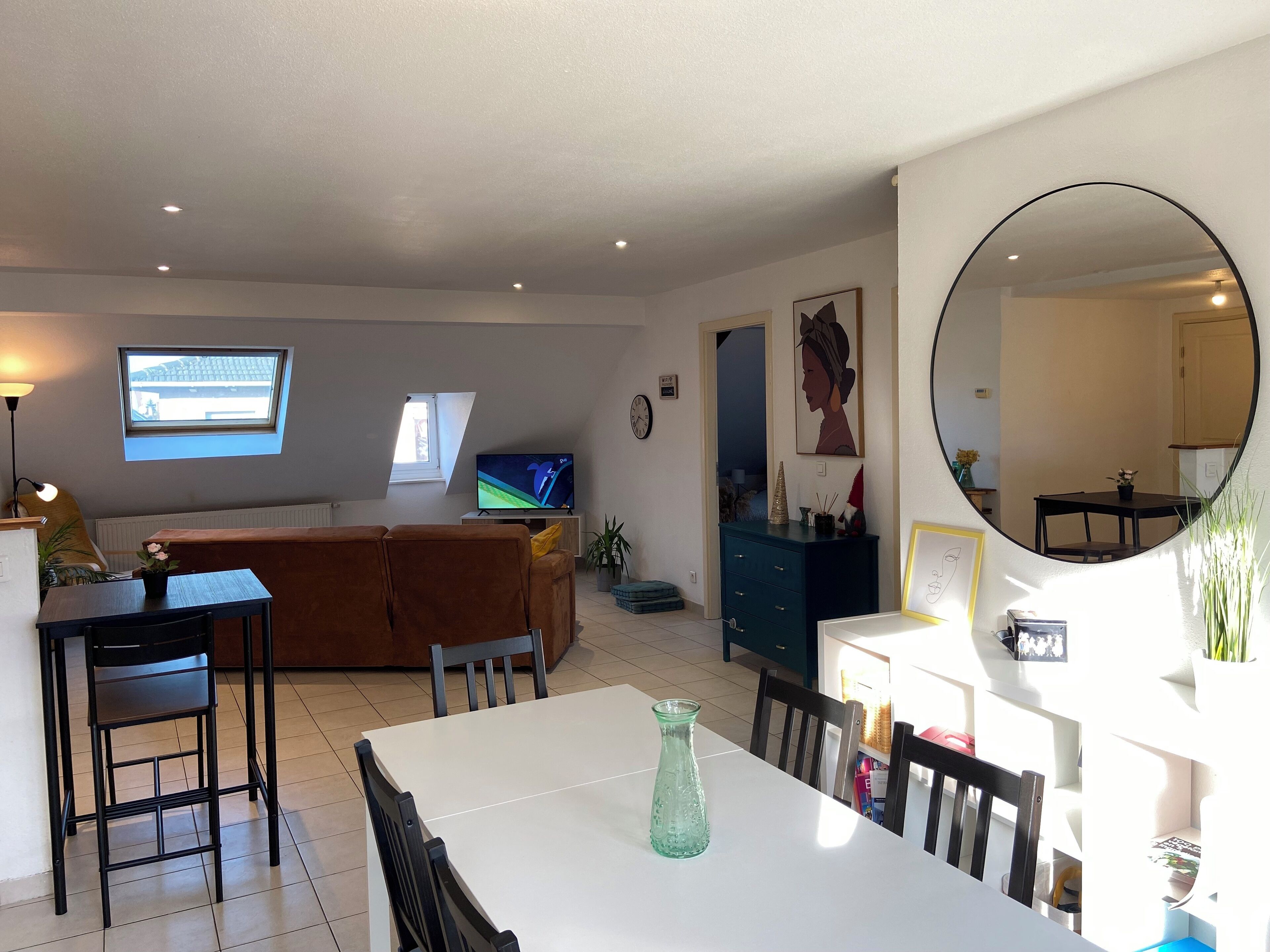 Een eethoek met een witte tafel, zwarte stoelen en een groene vaas. Er is een televisie op een dressoir, een raam met uitzicht en een spiegel aan de muur.