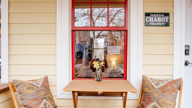 A wooden table with a vase of flowers, two chairs with patterned cushions, and a window with a reflection of a person and a lamp. - Cute 2-bed home, 3 blocks from downtown Bozeman, with a large yard! (Bozeman)