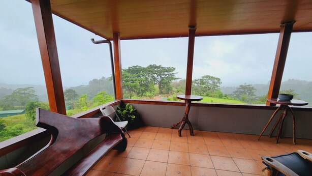 Un balcon avec un banc en bois, une petite table sur laquelle se trouve un bol, et une vue sur un paysage verdoyant.