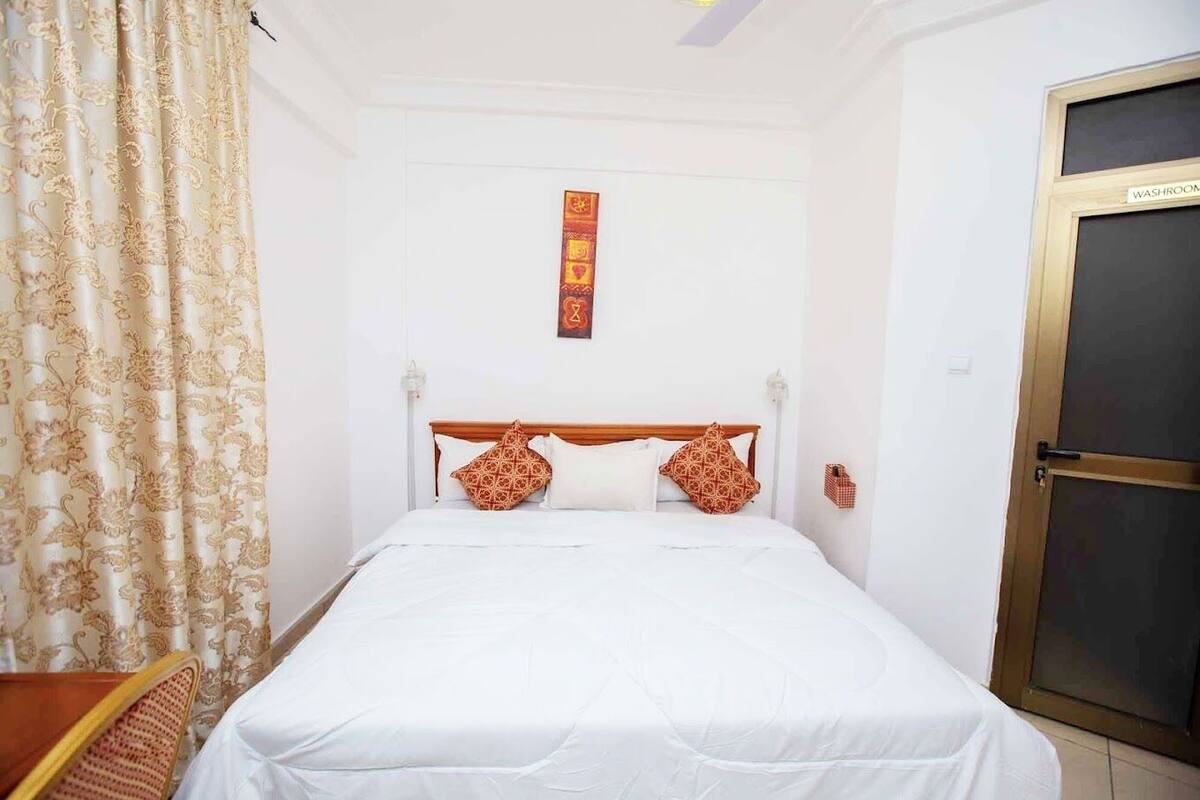 A neatly made bed with white bedding, a wooden headboard, and patterned pillows. A decorative wall hanging is visible above the bed.
