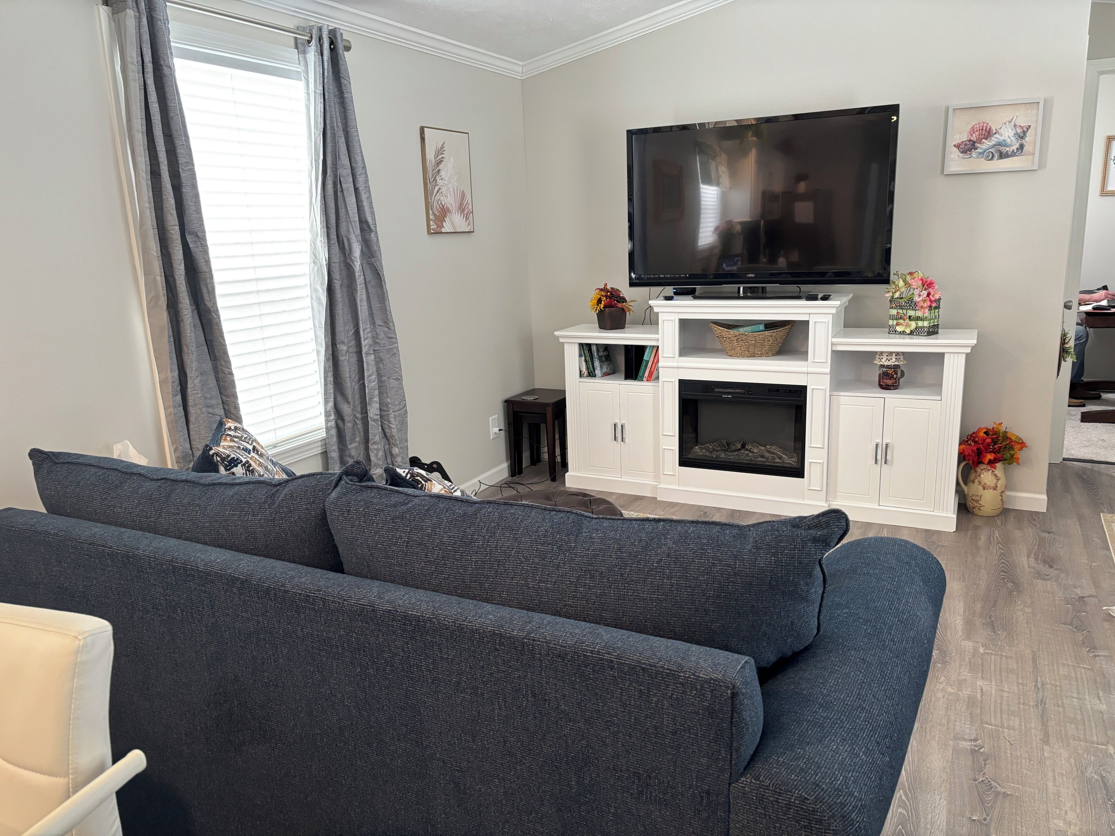 Ein Wohnzimmer mit grauem Sofa, einem Flachbildfernseher auf einer weißen Unterhaltungsanlage, einem Kamin und einem kleinen Tisch mit Dekoration.