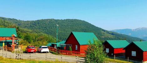 A row of red cabins with green roofs, a parking area with a red car, and a mountainous backdrop.