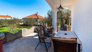 A covered outdoor seating area with a table and chairs, a stone wall, and a view of a house and trees. - Two Bedroom Apartment with Terrace (Ugljan)