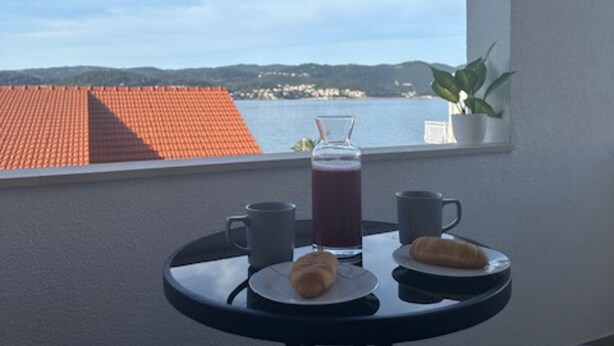 Un balcón con una mesa dispuesta para el desayuno, un vaso de jugo y vista a un pueblo costero.