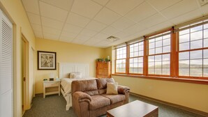 A room with a brown leather sofa, a wooden coffee table, a bed with a white headboard, a nightstand, and a window with a view of trees. - Day Trip to Pittsburgh: Comfy New Kensington Apt! (New Kensington)