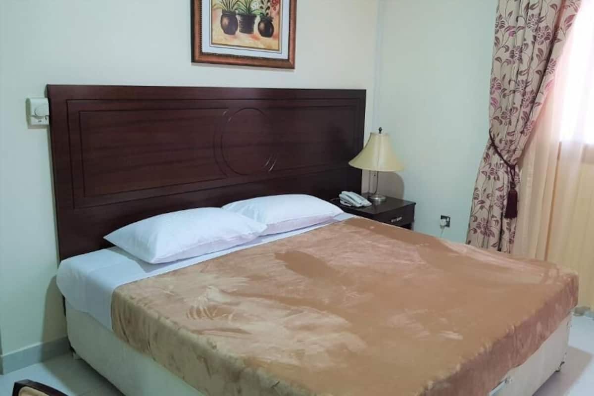 A neatly made bed with a brown bedspread, a wooden headboard, a bedside lamp, and a framed picture on the wall.