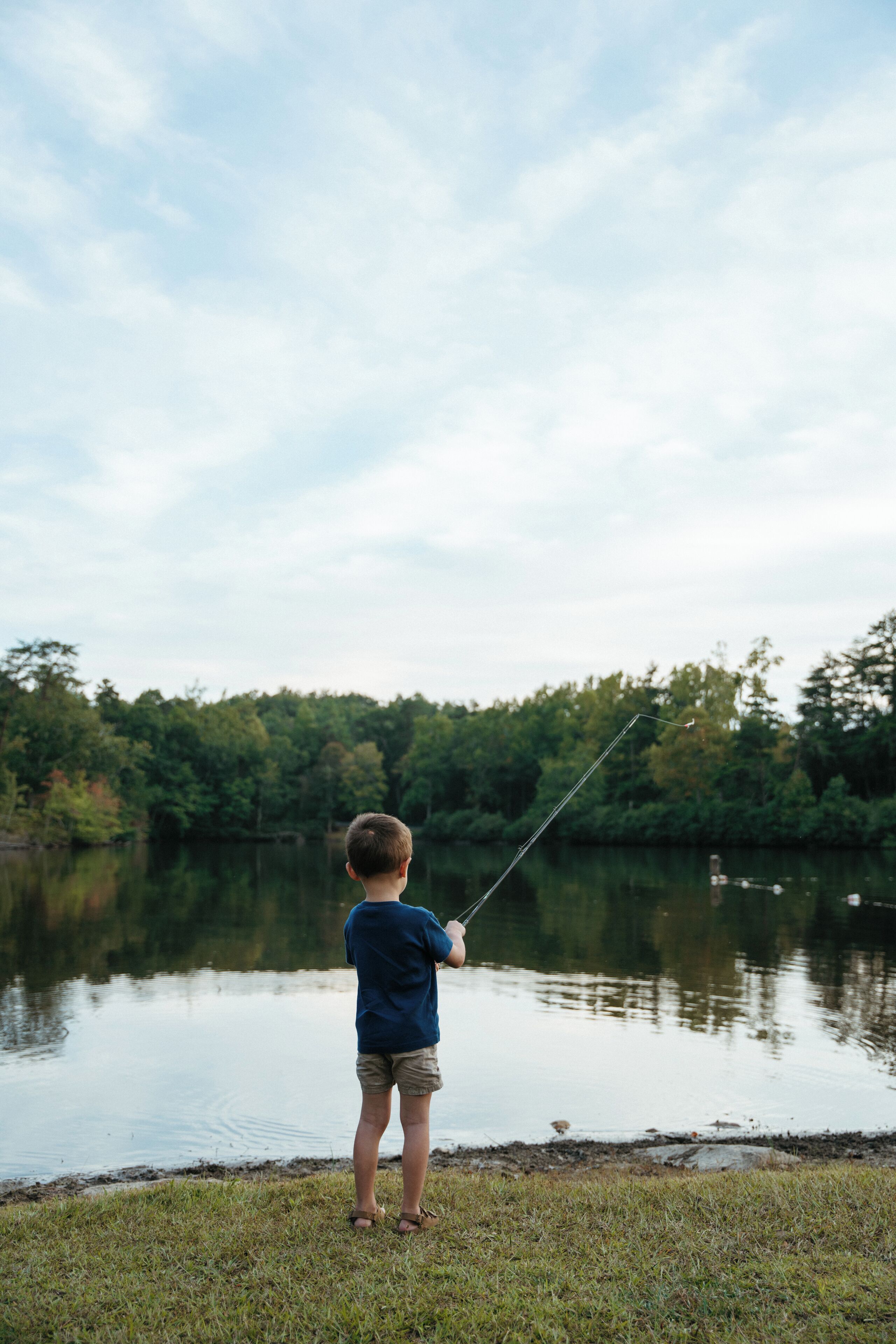 Un niño pescando en un lago tranquilo con un bosque al fondo.