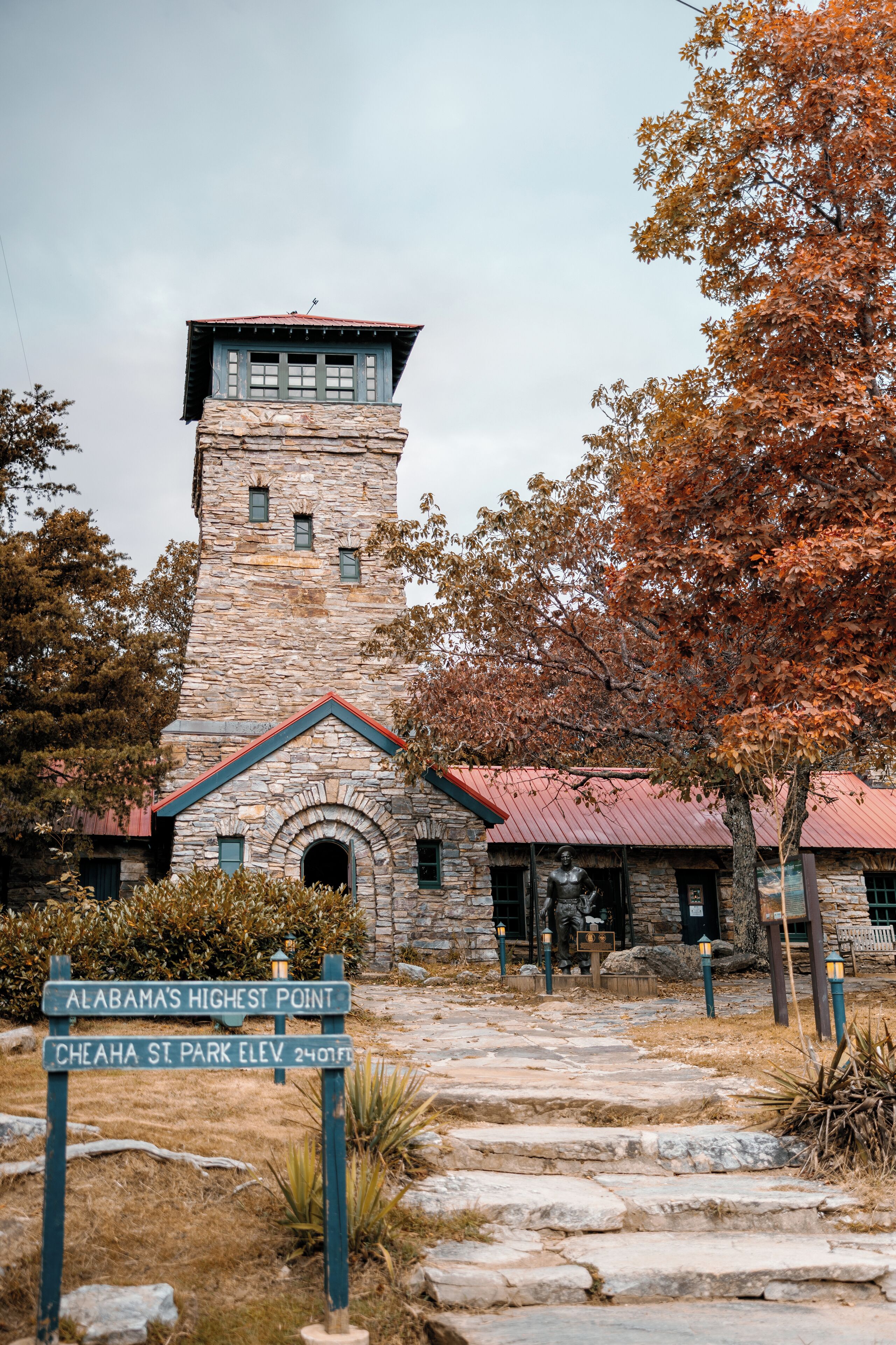 Un edificio in pietra con una torre, un cartello che indica 'Punto più alto dell'Alabama' e 'Parco statale di Cheaha, altitudine 2407 piedi', e un sentiero in pietra che conduce all'ingresso.