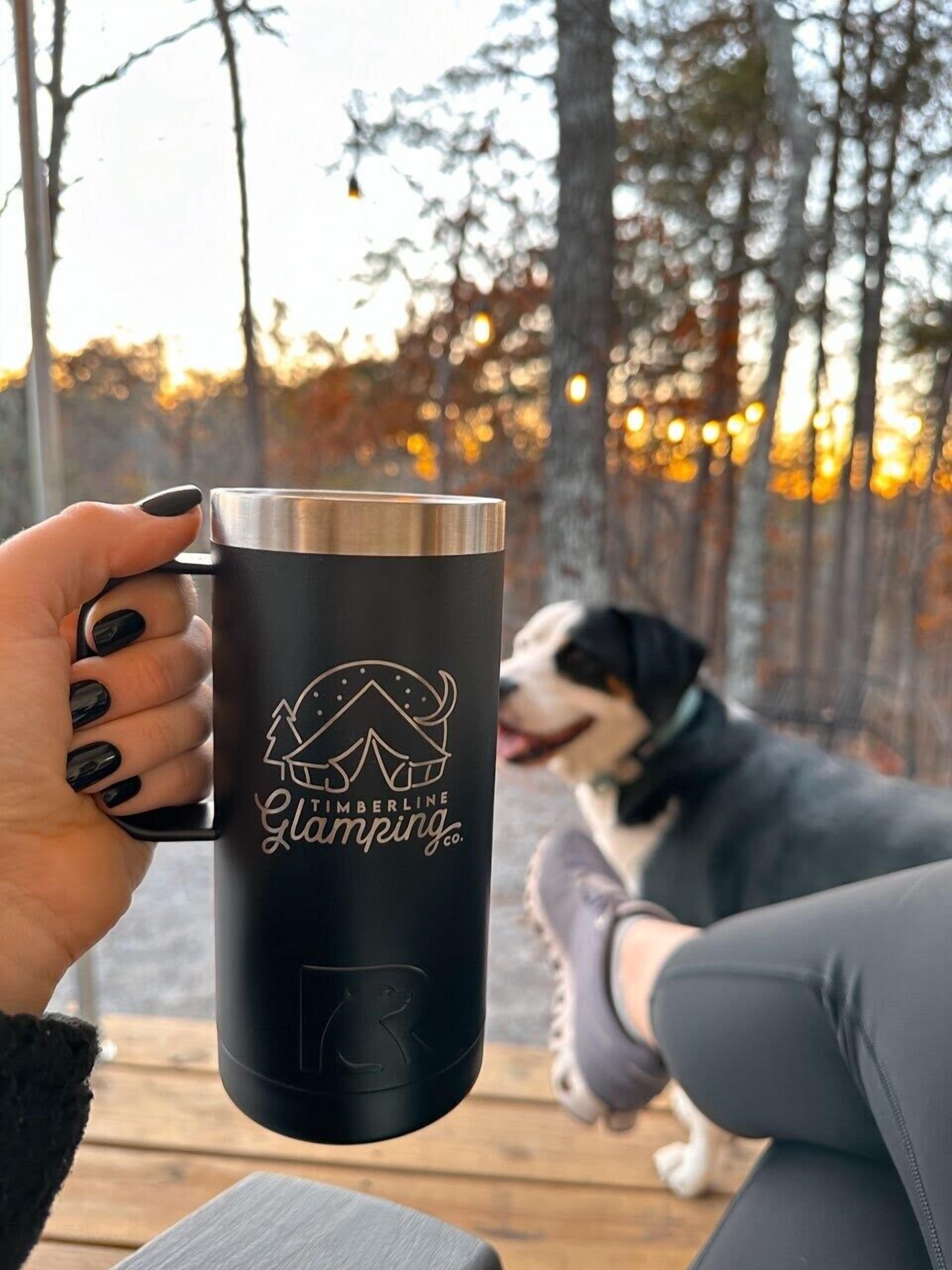 A person holding a black mug with a tent and 'Timberline Glamping' logo.
