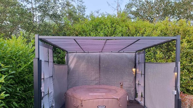 A hot tub in a covered outdoor area with a metal frame and mesh walls, surrounded by greenery. - Studio à Louer (Clohars-Carnoët)
