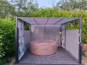 Een hottub in een overdekte buitenruimte met een metalen frame en gaaswanden, omgeven door groen.