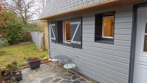 A house with a stone patio, two windows with shutters, and a white door. - Studio à Louer (Clohars-Carnoët)
