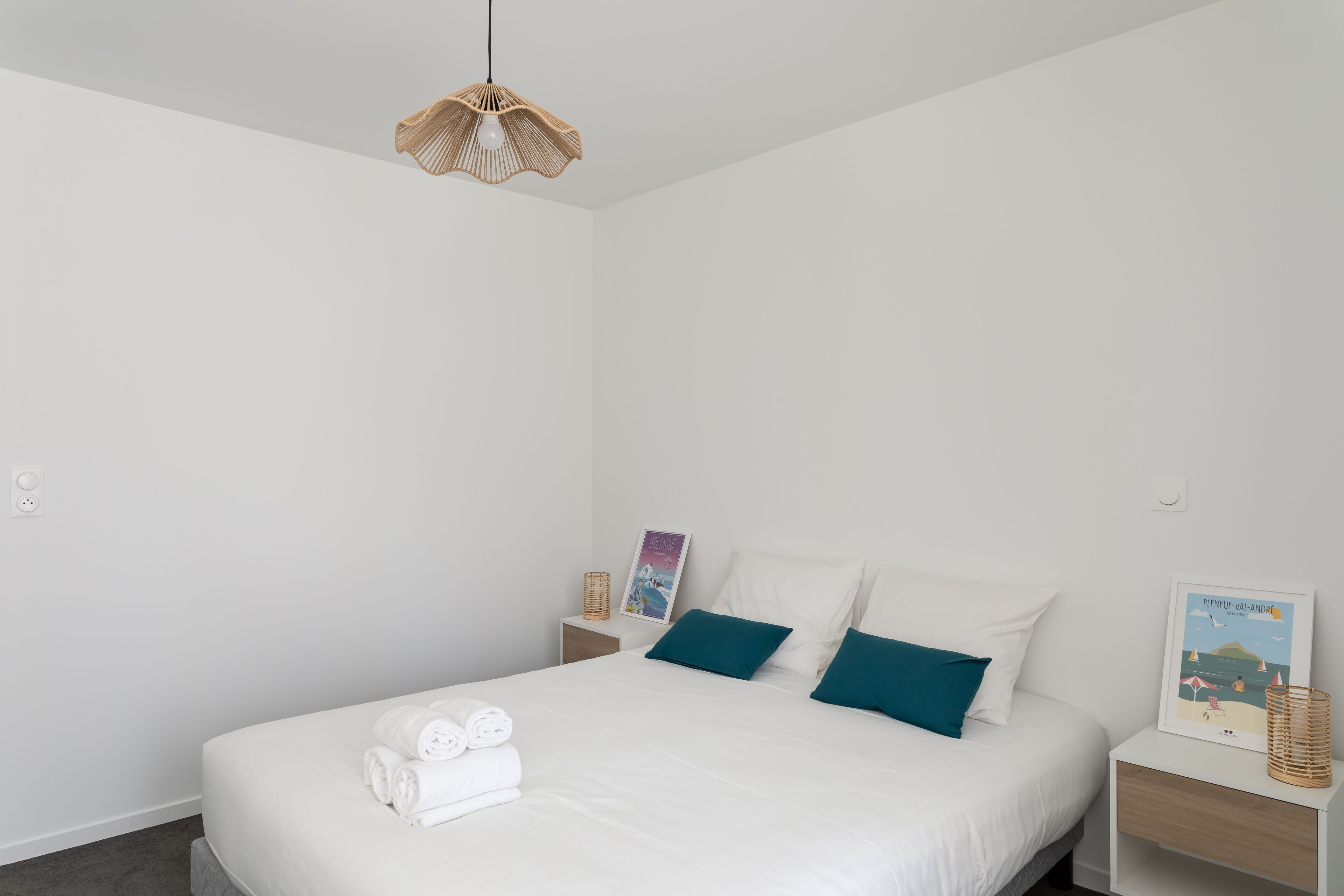 A neatly made bed with white linens and teal pillows, a hanging light fixture, and a small bedside table with a lamp and a framed picture.