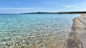 Ein sauberer Strand mit Kieselsteinen und ruhigem Wasser, ein Boot in der Ferne und eine hügelige Landschaft im Hintergrund.