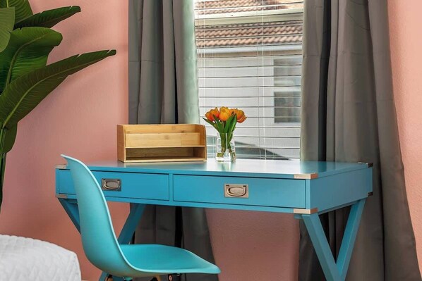 Una habitación con un escritorio azul, una silla, un ventanal con cortinas y una planta.