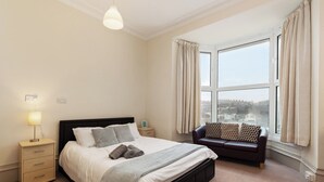 Quarto com cama, mesa de cabeceira, abajur e vista para a cidade pela janela.