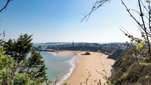 Vista costeira com uma praia de areia, céu claro e edifícios ao fundo.