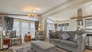 A spacious living room with a grey sofa, matching ottoman, and two armchairs. A chandelier hangs from the ceiling, and a kitchen is visible in the background. - Cormorant - Luxury Lodge, Close to the Beach (Tenby)
