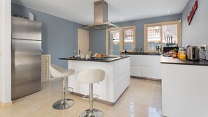 A modern kitchen with a stainless steel refrigerator, white bar stools, a central island, and a large window. - Villa Hilary with private pool near Playa de Muro (Muro)